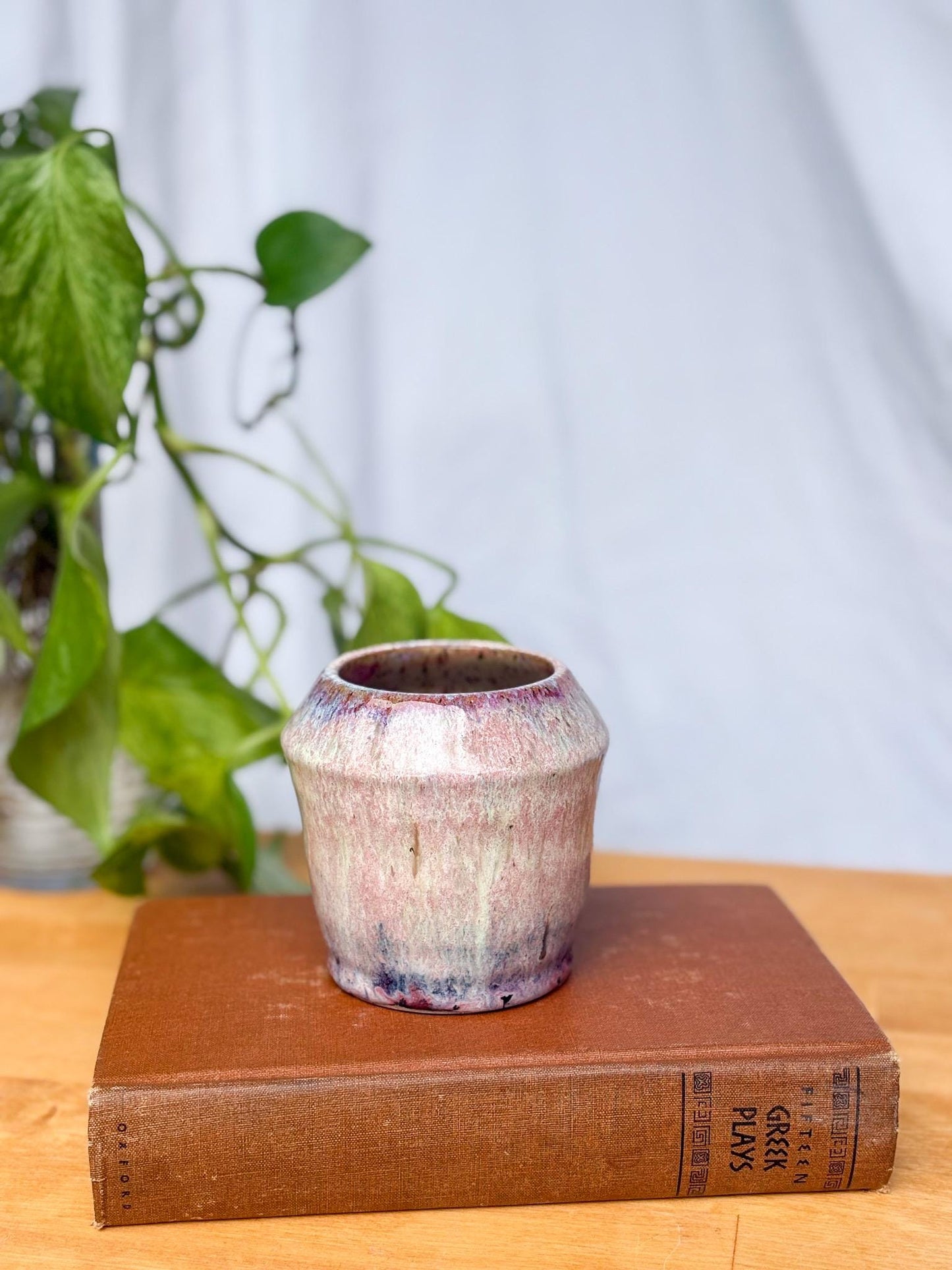 May include: A small, white ceramic planter with a purple and pink glaze. The planter has a rounded bottom and a slightly flared top. It is sitting on top of a brown book with the title 'Fifteen Greek Plays' printed on the spine.