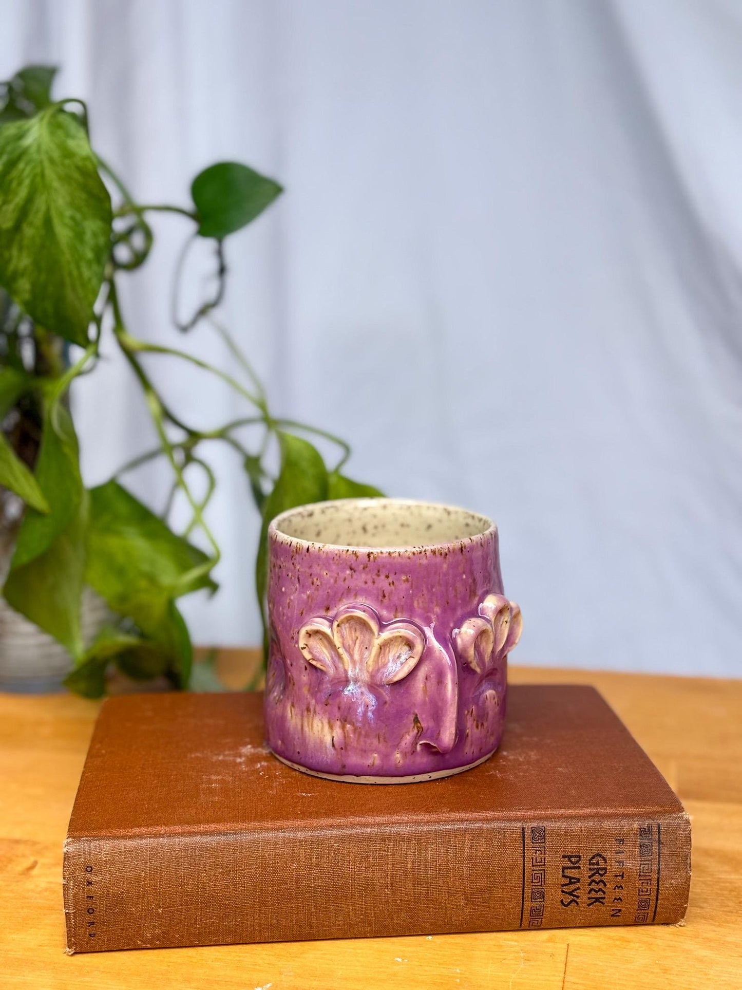 May include: A purple ceramic mug with a textured surface and three raised flower-shaped details. The mug is sitting on top of a brown book with the title &#39;FIFTEEN GREEK PLAYS&#39; printed on the spine.